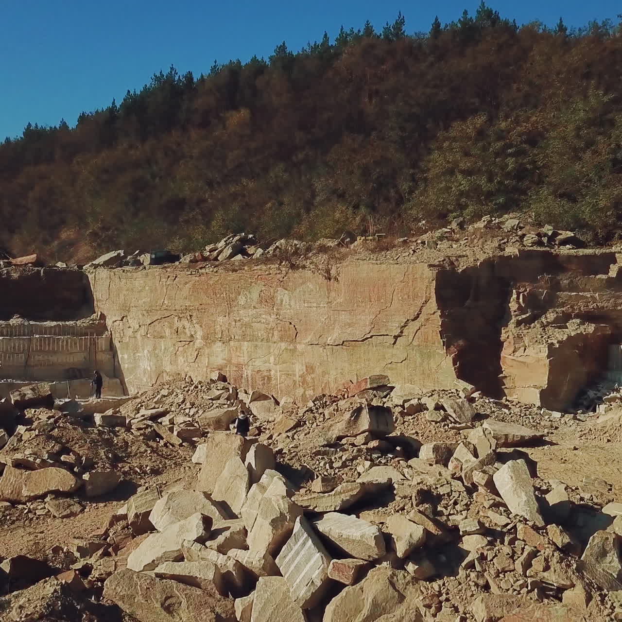 People are working around heaps of huge stones near a high and long quarry with stone rocks and with a coniferous forest on top in a warm summer day on the background of blue sky. Aerial view. Camera motion to forward.