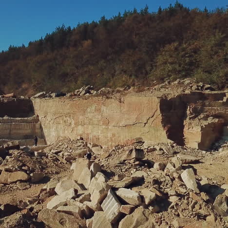 people are working around heaps of huge stones near a high and long quarry with stone rocks and with a coniferous forest on top in a warm summer day on the background of blue sky. aerial view. camera motion to forward.
