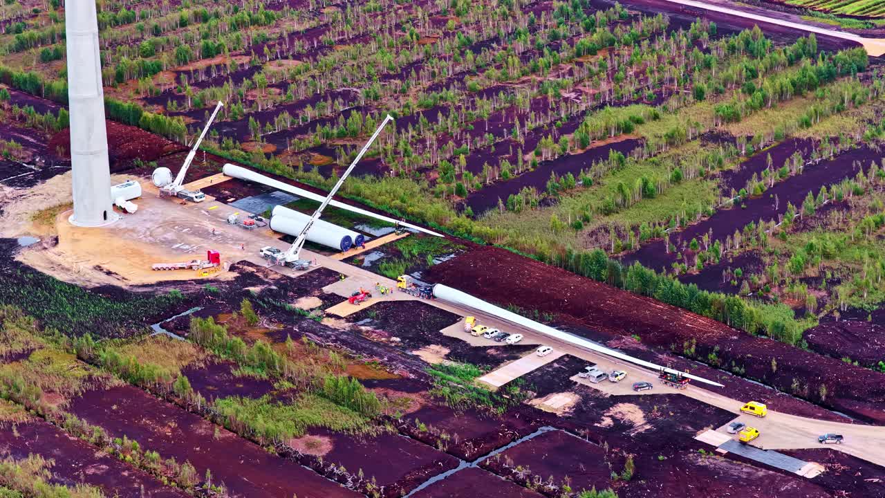 Cranes and equipment assemble wind turbine parts beside a tall tower in agricultural fields