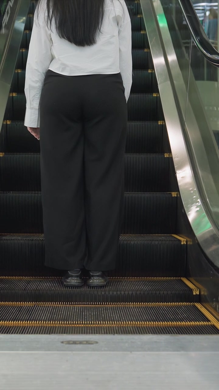 Primer plano de una escalera mecánica subiendo. La cámara capta los escalones vacíos subiendo y bajando, enfocando. El fondo presenta paredes de cristal que reflejan el espacio interior con la escalera mecánica.