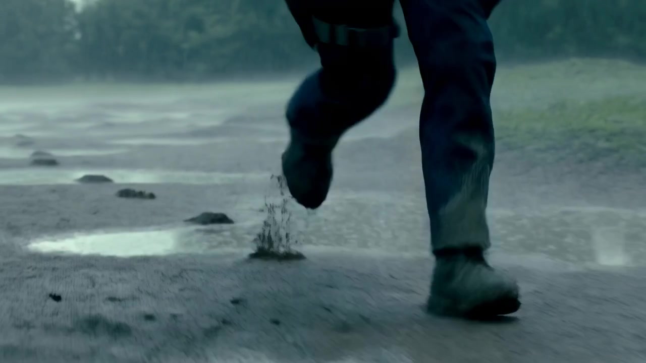 Primo piano delle gambe e degli stivali di una persona che si muovono sulla sabbia umida accanto all'acqua bassa, con schizzi, nebbia e un'atmosfera fredda e piovosa che crea un'immagine suggestiva.