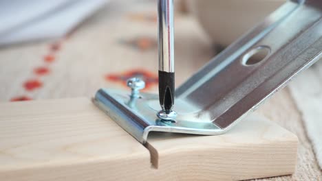 close-up of a person's hand using a screwdriver to screw a screw into a metal bracket attached to a wooden board.