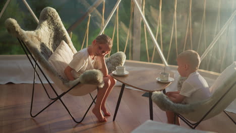 sleepy children rest sitting at table in early morning. little boy and girl drink tea in tent at resort in nature reserve. summer family vacation