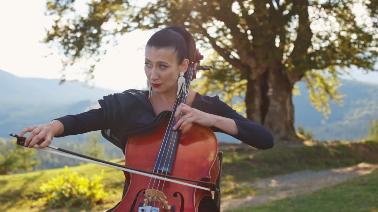 Violoncello musician with bright makeup and long earrings playing instrument. Lady stops to fix the earring and goes back to playing. Nature backdrop.