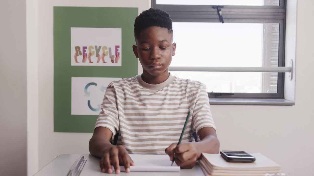 In school, African American teenage boy writing in notebook at desk with smartphone and books