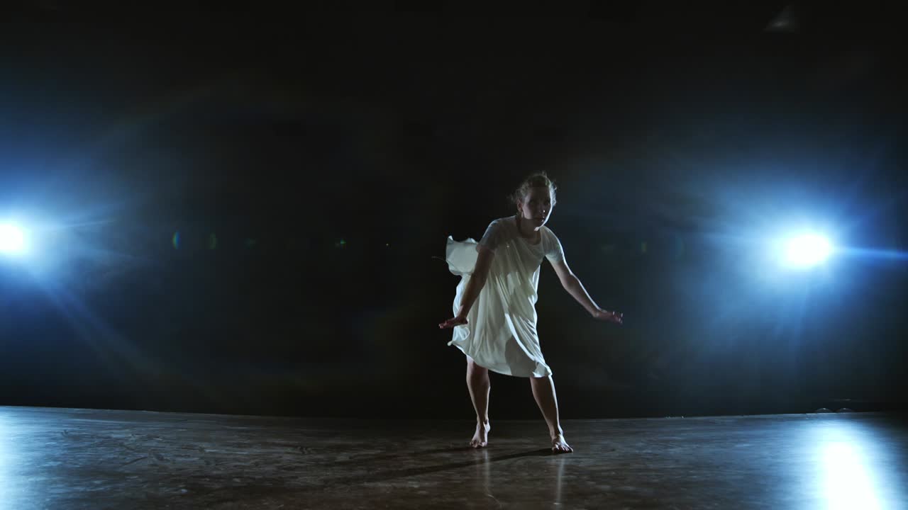 A young female ballerina barefoot jumps on stage and moves in slow motion in a loose white dress