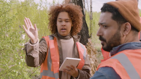 Close-up-view-of-an-african-american-woman-activist-holding-a-tablet-and-talking-with-her-coworkers-in-the-forest-while-they-deciding-where-to-plant-trees
