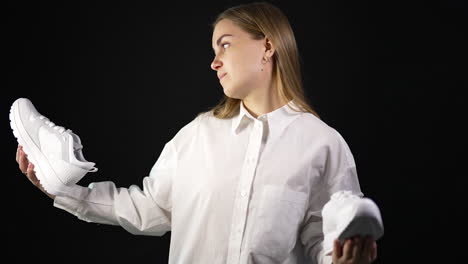 long-haired blonde lady in white shirt holding white sneakers in both hands. young woman making choice between two types of footwear. black backdrop.