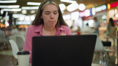 mujer de vestido rosa trabajando en una computadora portátil mientras come una hamburguesa en una mesa en un centro comercial ocupado, una taza de café está colocada al lado de la computadora portatil, y ella está enfocada en su tarea, multitarea entre el trabajo y la comida