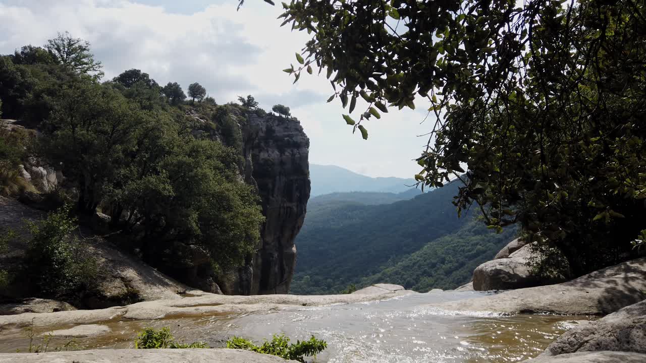 Vista panoramica di un ruscello che scorre sulle rocce verso il bordo di una cascata, con una vista mozzafiato sulla valle verde e le montagne a Rupit, Catalogna, in una giornata di sole.