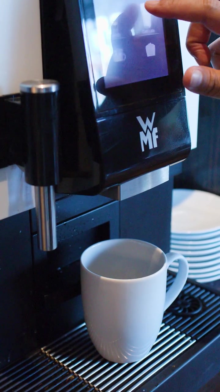Primo piano di una mano che posiziona una tazza bianca e seleziona il caffè su un touchscreen.