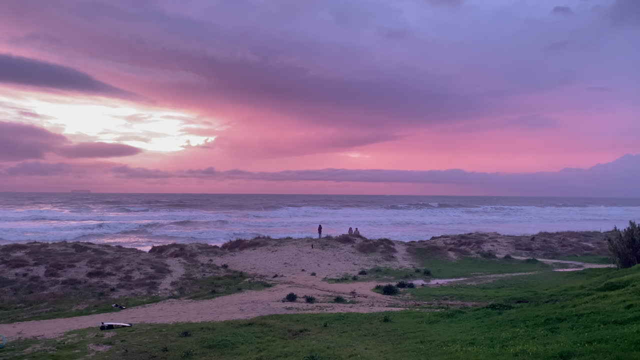 Persone che si godono un bellissimo tramonto sulla spiaggia di Cadice, sentendosi in pace e calmi mentre osservano nuvole rosa e viola drammatiche sulle onde dell'oceano.