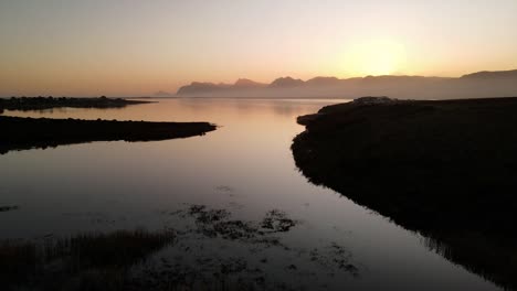 Vuelo-Calmante-Sobre-El-Estuario-De-Una-Laguna-Mientras-El-Sol-Se-Pone-Con-Las-Montañas-En-La-Distancia