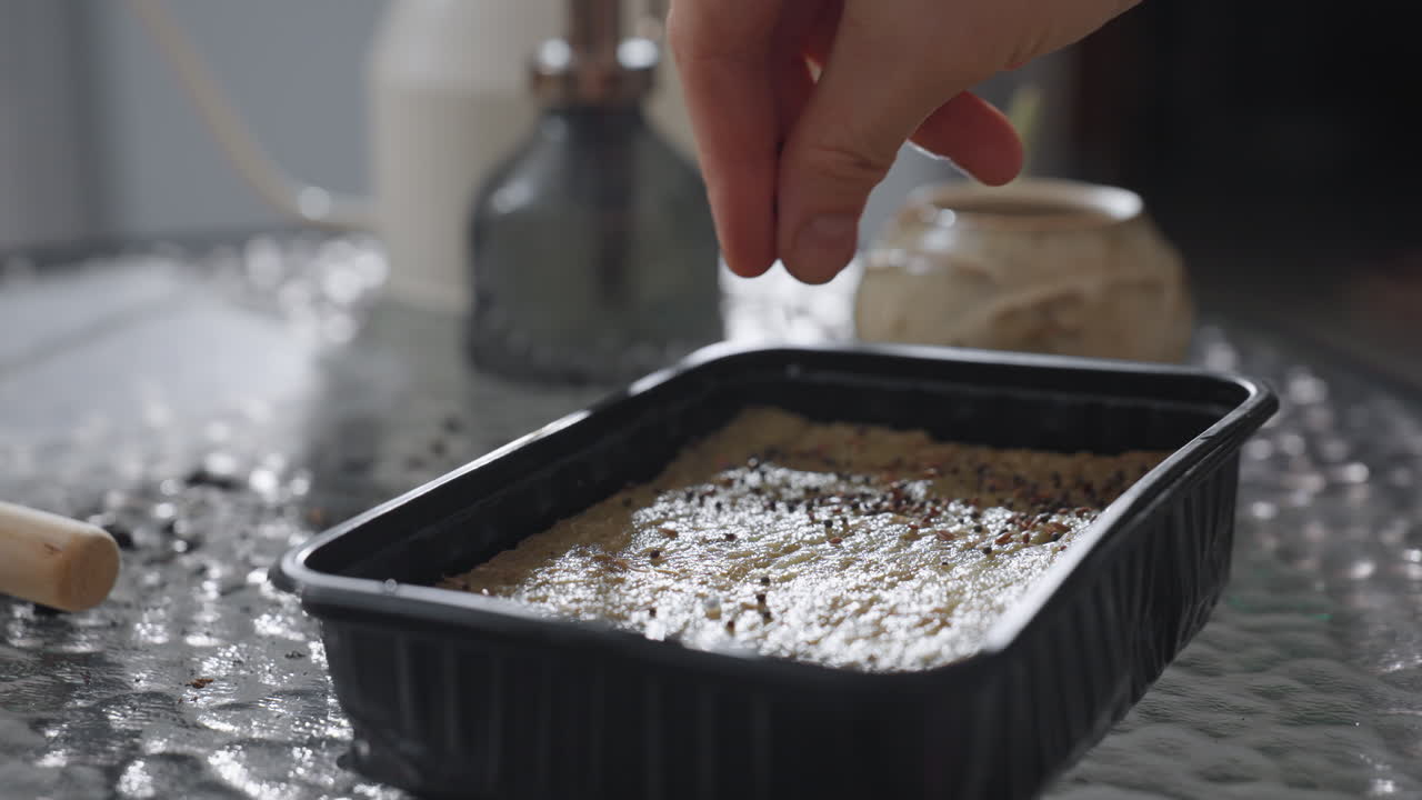 Close up of green thumb enthusiast carefully sprinkling tiny seeds onto moist soil in black tray on glass table with watering can, ceramic planters, indoor gardening tools capturing seed planting