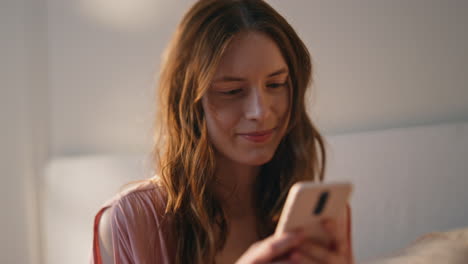 Girl texting mobile phone in bed closeup. Smiling young woman typing smartphone