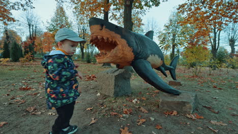 a life-size wooden sculpture of a shark in the park. baby boy comes up to the sculpture and puts some chestnut into the jaws.