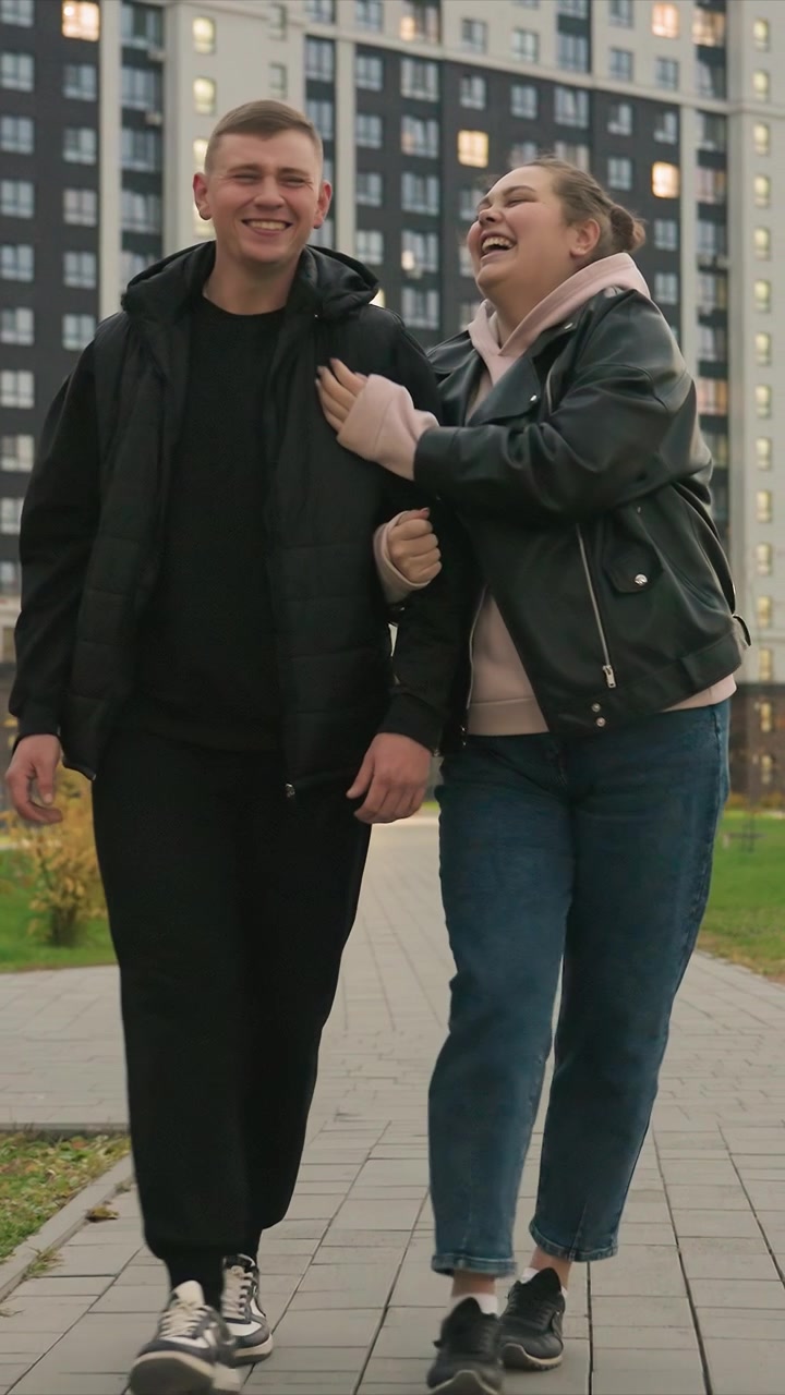 Gli individui procedono insieme tenendosi per mano. La coppia mostra un movimento delle gambe coordinato mentre camminano. La scena presenta un sentiero pavimentato all'aperto fiancheggiato da vegetazione e residenze a più piani.