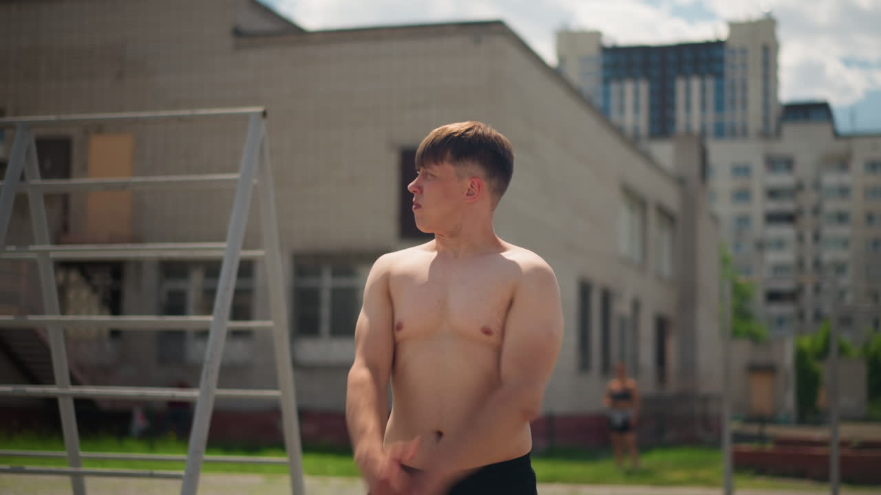 상의를 탈의한 운동선수가 도시 안뜰 환경에서 조정 능력을 향상시키고 있습니다. 백인 혈통의 운동선수가 훈련된 집중력으로 도시 환경 속에서 야외 안정성 운동을 하고 있습니다.