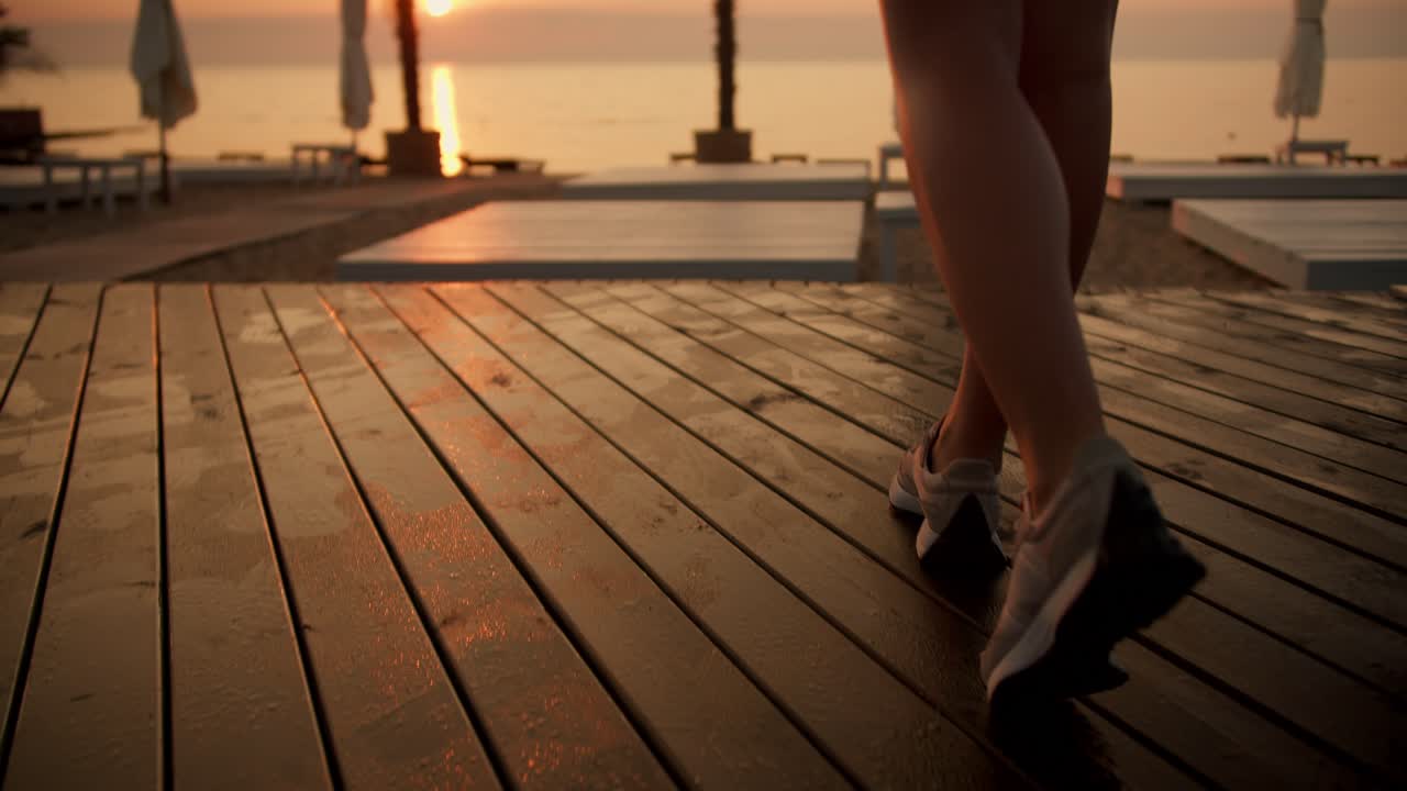 En pige i en sort sportssommeruniform går langs stranden med brætter og drikker vand fra en sportsflaske ved daggry. Forbereder sig til sportsaktiviteter om morgenen, meditation ved solopgang