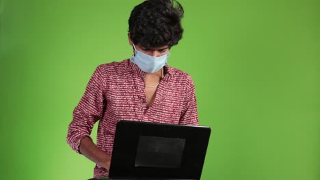 joven hombre de negocios feliz en una camisa roja con máscara trabaja en una computadora portátil en una pantalla verde