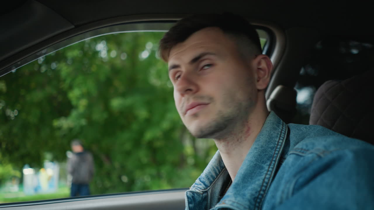 Ung mann som kjører bil, ettertenksom stemning, nærbilde av denimjakke, interiørportrett med grønne trær synlig gjennom vinduet, naturlig, mykt lys, hender på rattet, venter ved veikanten, filmatisk