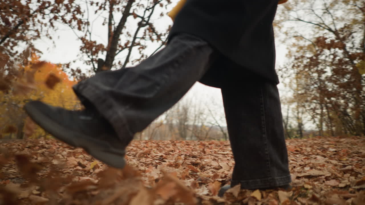 Vista delle gambe di un individuo che cammina attraverso la foresta con fogliame autunnale secco, calciando e disperdendo giocosamente le foglie, indossando scarpe di tela e jeans neri, creando un effetto di movimento dinamico nella natura