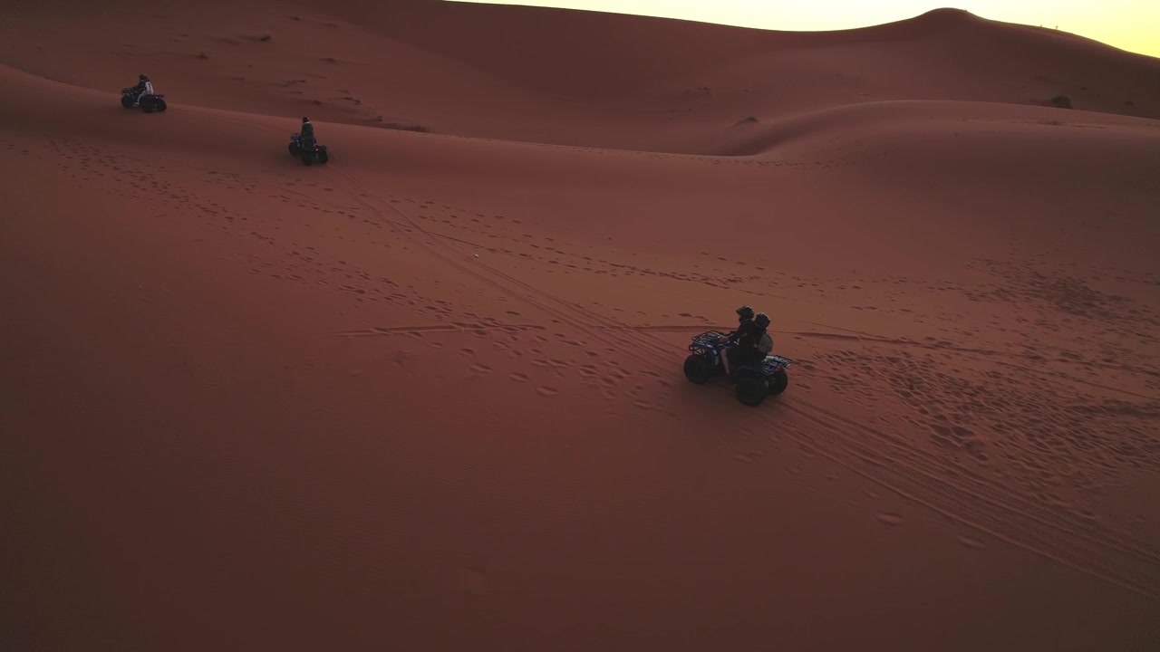 Quad che attraversano le dune dell'Erg Chebbi vicino a Merzouga, Errachidia, Drâa-Tafilalet, Marocco, Africa, lasciando tracce nel Sahara durante la calda luce del tramonto, ripresa di tracciamento panoramica con drone.