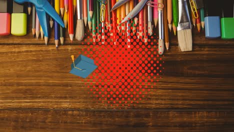 Animation-of-mortarboard-and-textbook-bouncing-on-red-dots-over-desk-with-pencils-and-brushes