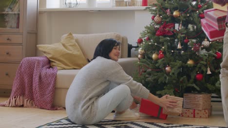 brunette woman sitting under christmas tree, her husband brings her christmas gifts