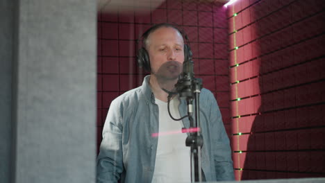 a man wearing a blue long sleeve shirt, white undershirt, and black headphones is singing passionately in a recording studio. he stands in front of a microphone with a red acoustic panel