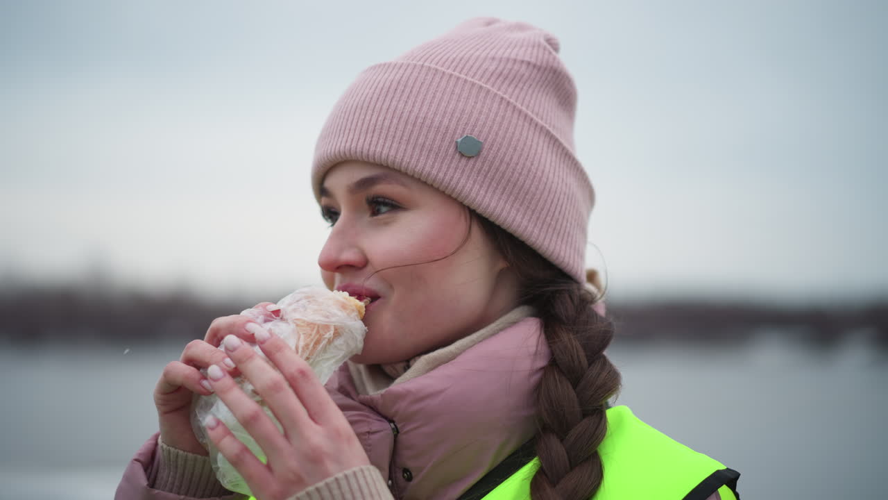 Young woman in pink winter jacket and beanie wearing reflective safety vest standing outdoors near water on cold overcast day, looking ahead with calm and thoughtful expression during work
