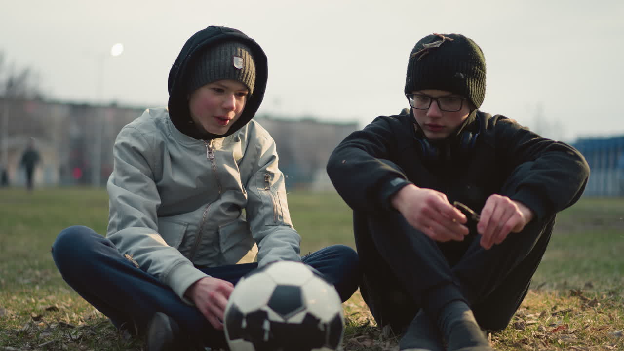 Two boys seated on a grassy field, engaged in conversation, one boy, wearing glasses, picks a piece of grass from the ground and breaks it, while grass particles are seen flying around