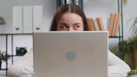 Confident young business woman hiding behind laptop computer looking at camera spying peeping