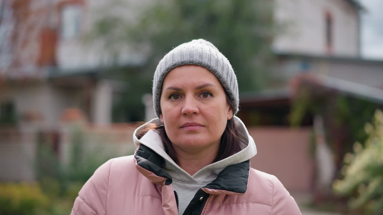 분홍색 재과 회색 베니를 입은 자신감 넘치는 여성이 차분한 표정으로 카메라를 쳐다보고, 뒷면에 한 가을 잎자루, 집, 정원과 함께 야외에 서 있습니다.