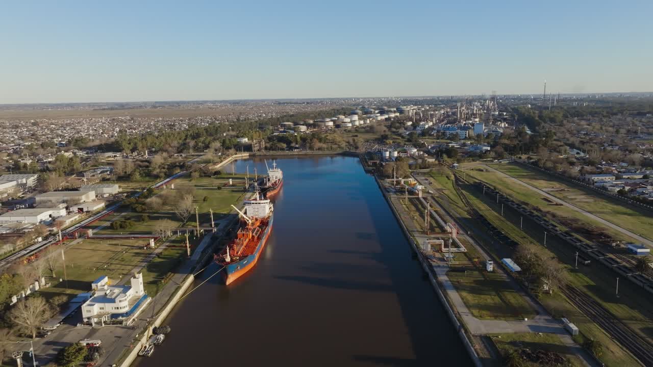 Daytime drone view of a cargo ship navigating a river port with docks, cranes, and industrial facilities visible