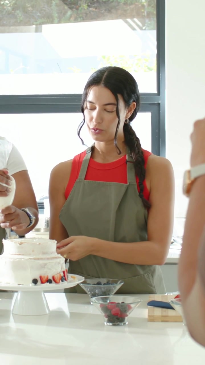 Video verticale: Una donna in maglietta rossa, con una sac à poche in mano, guida gli altri mentre tiene una torta all'isola della cucina.