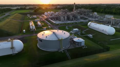 aerial view of industrial facility at sunset: tanks, pipelines, and production units surrounded by lush greenery and horizon glowing in evening light