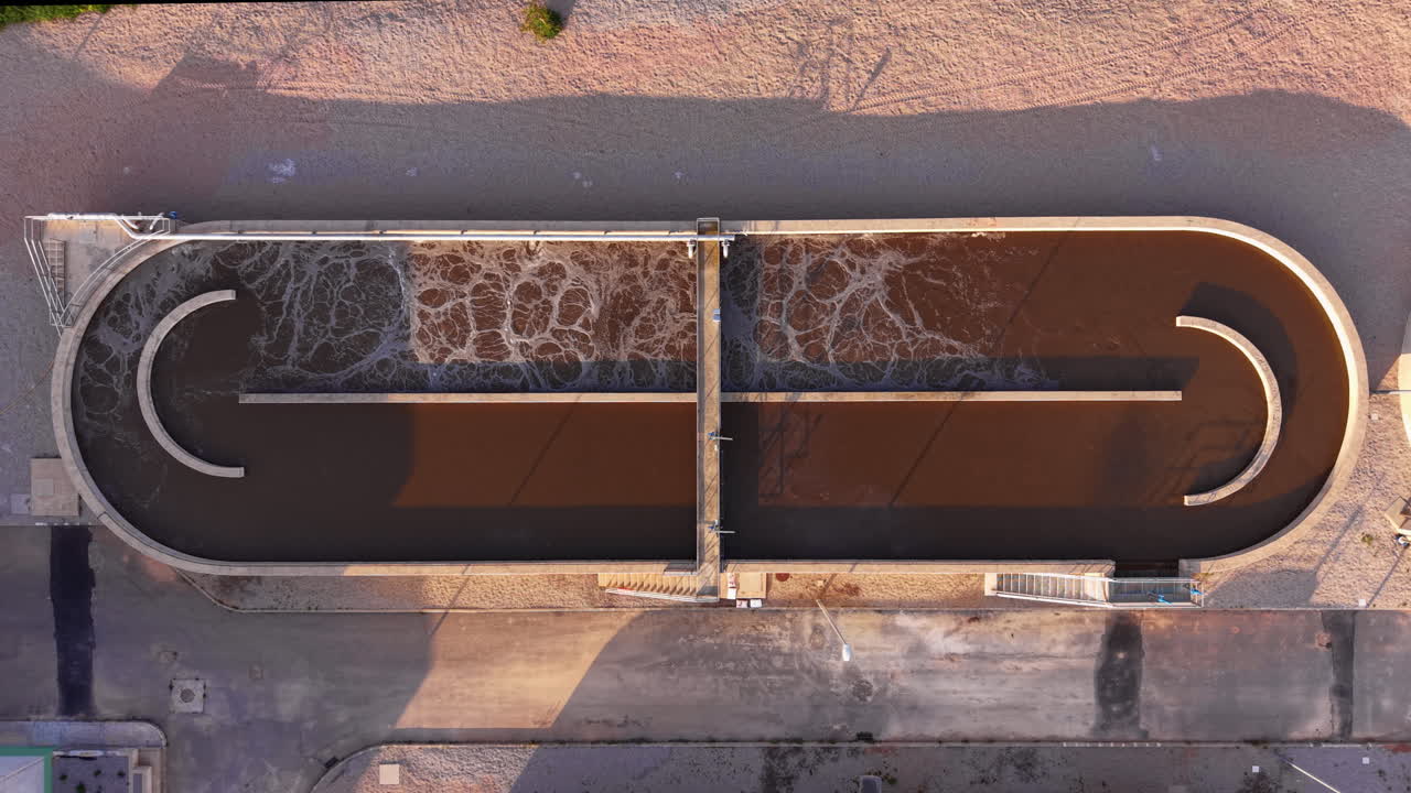 Top-down view of an oval aeration basin at a wastewater treatment plant with active surface agitation and brown activated sludge