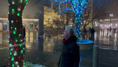 senior woman walking in a city decorated with christmas lights and night as is snows