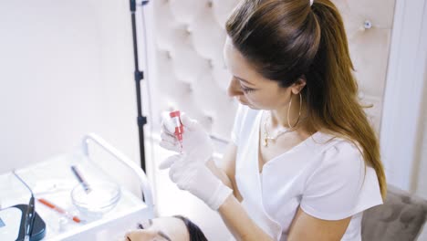 pretty beautician taking red ink to make permanent make-up in salon. cosmetologist in protective white gloves preparing equipment to make micropigmentation on lips for a young girl.