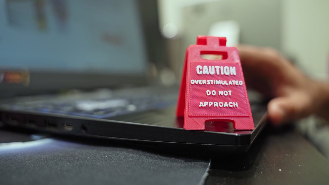 A red miniature caution sign warning of overstimulation sits on a black laptop keyboard. Close-up shot representing workplace stress and mental health awareness in a modern office setting.