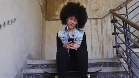 African-american-woman-wearing-earphones-using-listening-to-music-while-sitting-on-the-stairs