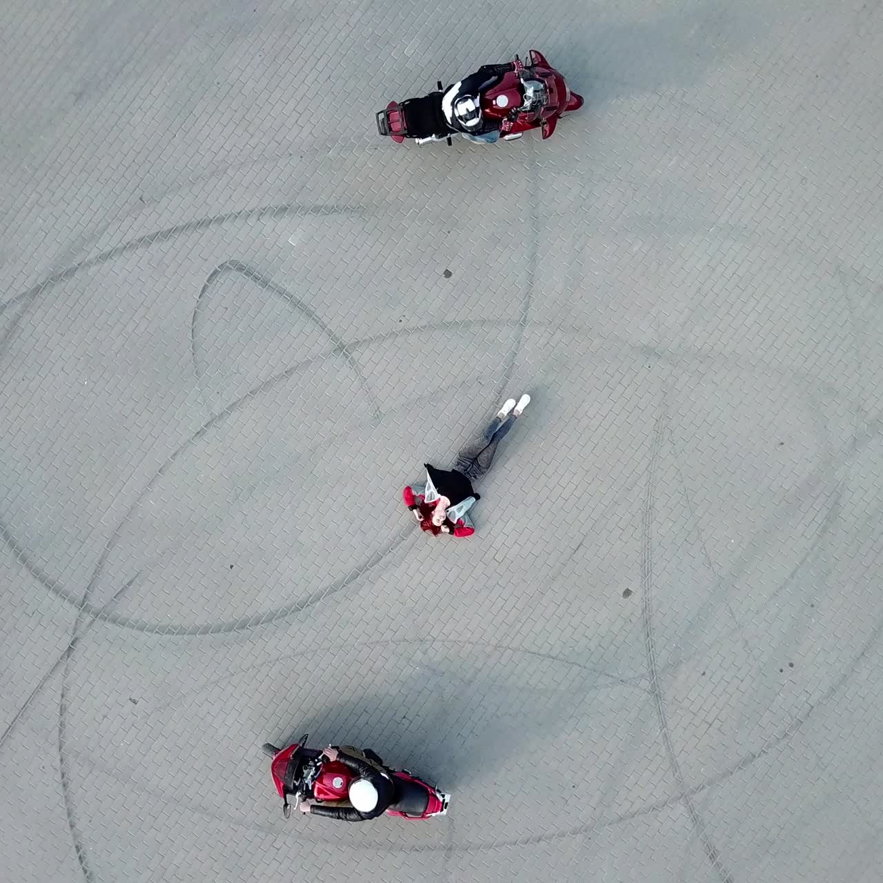 red-haired woman in sportswear is laying in the middle of pavement and surrounded by two motorcycles that are riding around her. Camera motion up
