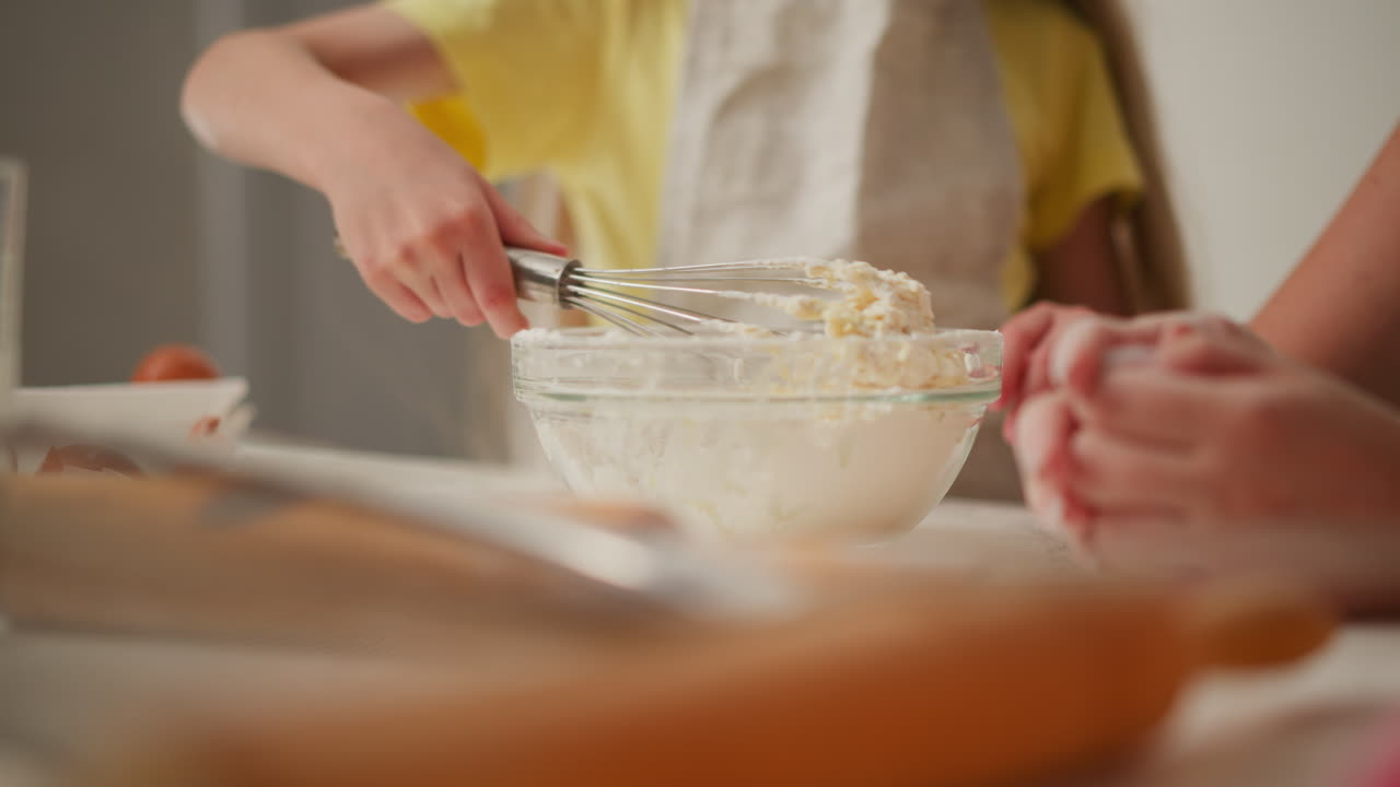 close up of hand using metal whisk to mix flour and egg dough in transparent bowl with cracked eggshells and kitchen tools visible in soft focus foreground, person sits beside observing process