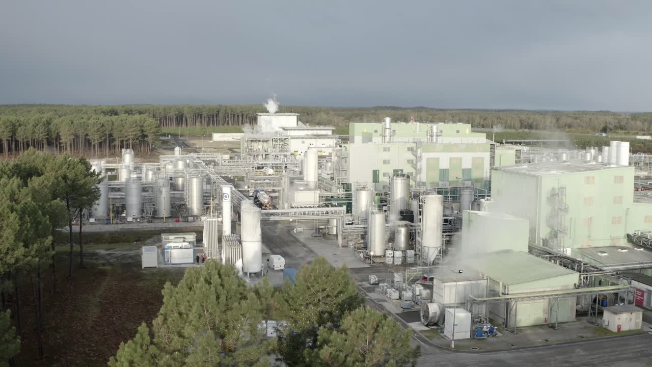 Drone footage flying over large chemical processing factory, numerous white storage tanks and buildings, steam rising, Castets, France