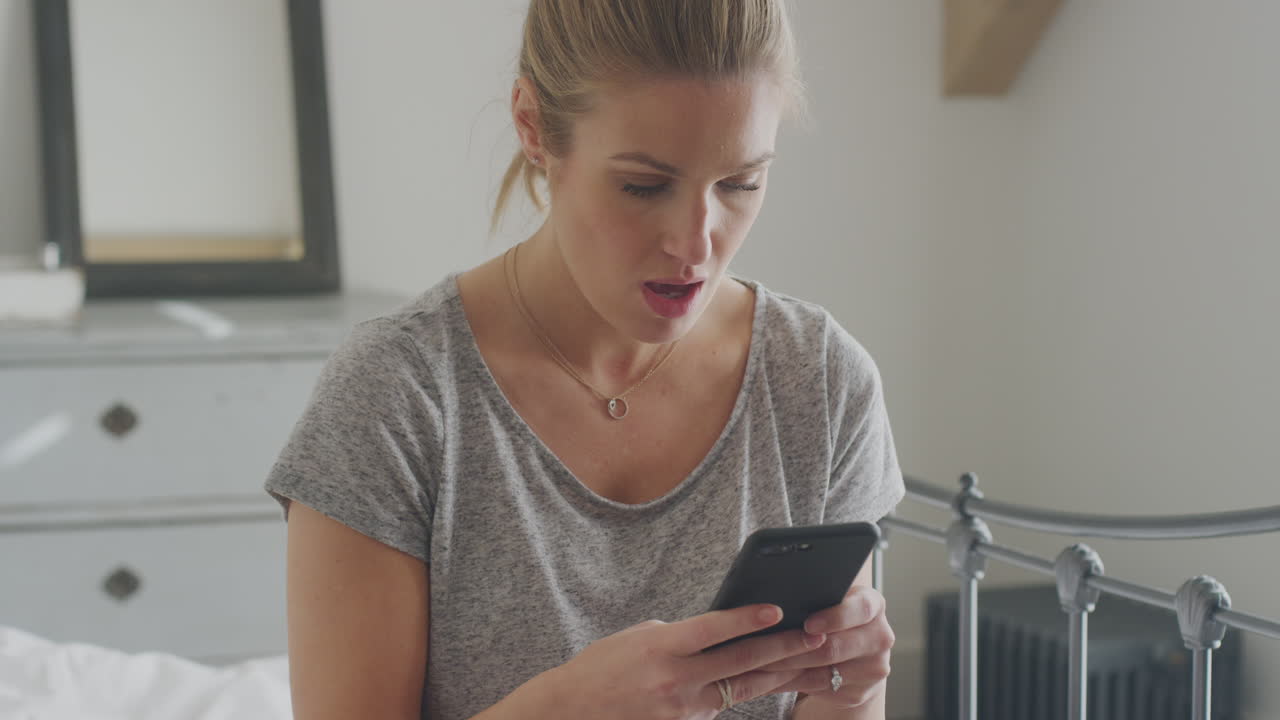 Unhappy Woman Wearing Pyjamas Sitting On Bed With Mobile Phone Being Bullied Online Via Social Media