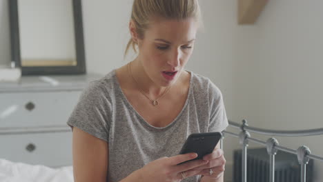 unhappy woman wearing pyjamas sitting on bed with mobile phone being bullied online via social media