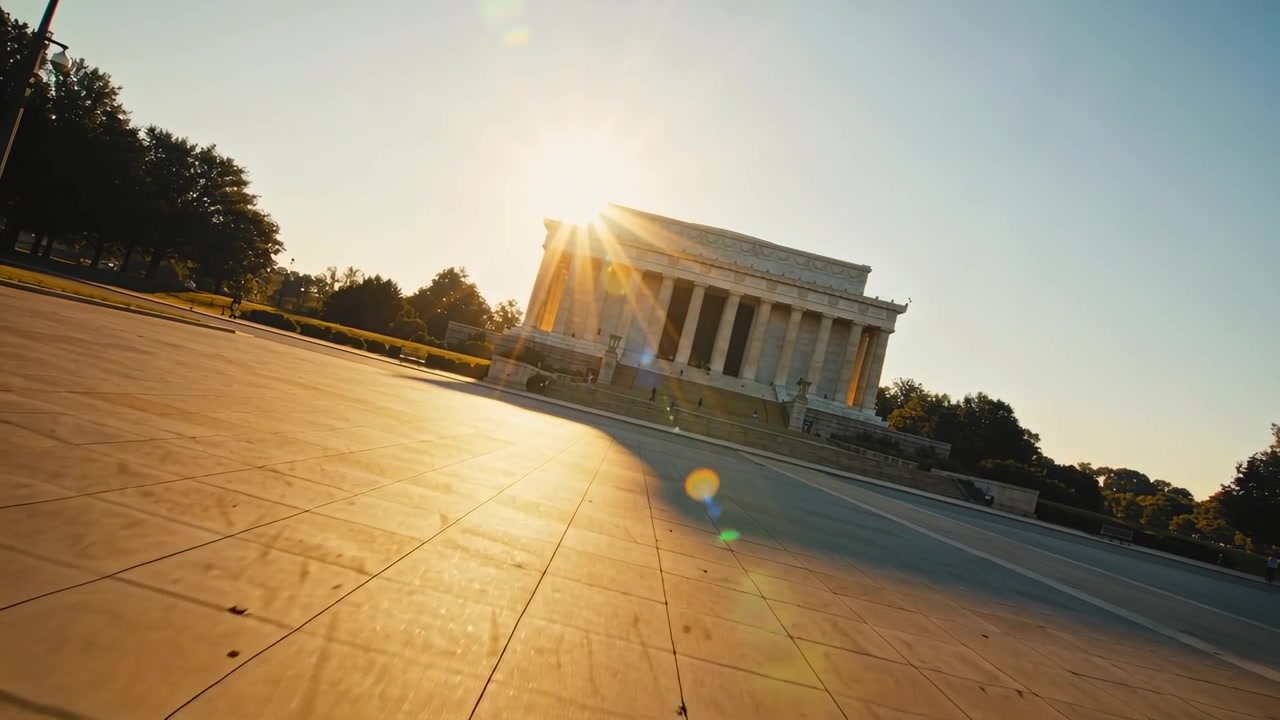 Telecamera che si inclina e si muove verso un monumento a colonne centrale su una piazza, catturando un bagliore solare e visitatori occasionali.