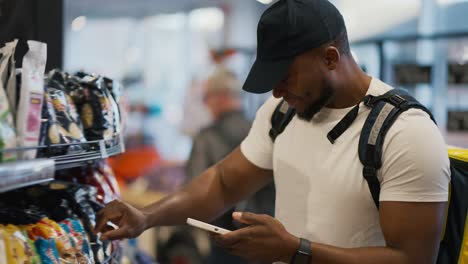 a happy and confident man with black skin in a white t-shirt and a black cap a food delivery person chooses the necessary goods and checks the list while holding a phone in his hands and a large yellow bag on his shoulders