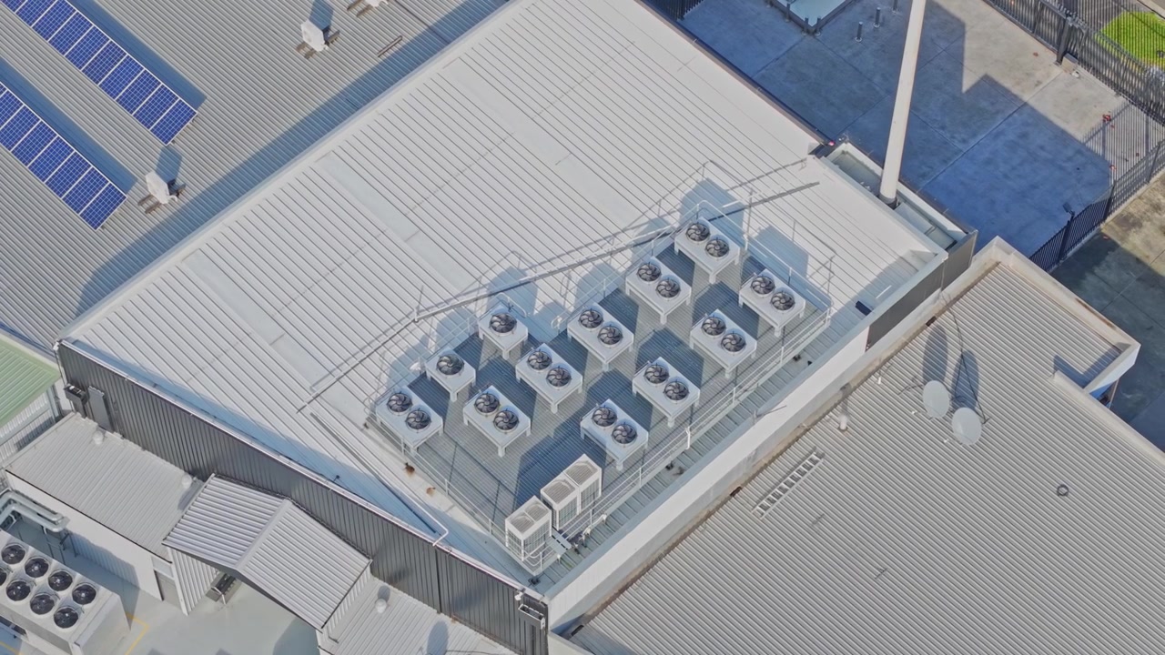 Drone captures a bright, sunlit rooftop of a data centre featuring industrial HVAC units, solar panels, and geometric metallic structures, highlighting energy and cooling technologies.